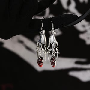 May include: Silver dangle earrings featuring a hand holding a cross and a red heart charm.