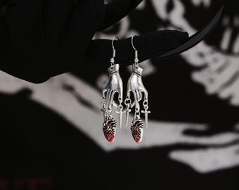 Puede incluir: Pendientes de plata con un diseño de mano sosteniendo una cruz y un dije de corazón rojo.