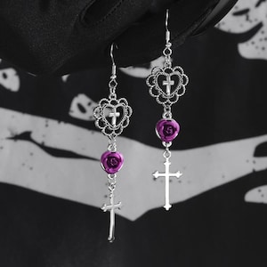 Puede incluir: Un par de pendientes de plata con un diseño de cruz, corazón y rosa. La rosa es de color morado oscuro.