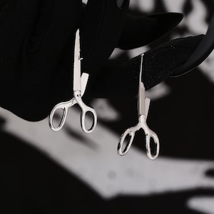 May include: Two silver scissor earrings. The earrings are hanging from a black surface.