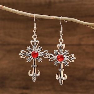 Puede incluir: Un par de pendientes de plata con colgantes de cruz de cristal rojo. Las cruces tienen detalles intrincados y un diseño de flor de lis en la parte inferior.