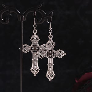 May include: A pair of silver cross earrings with intricate details. The crosses are suspended from silver hooks.