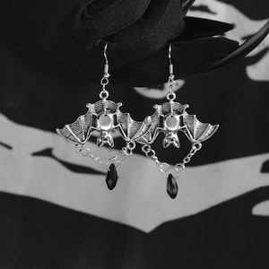 Könnte beinhalten: Silberne Fledermaus-Ohrringe mit detaillierten Flügeln und einem schwarzen, tropfenförmigen Anhänger. Die Ohrringe sind an einem Haken und einer Kette befestigt. Der Hintergrund ist schwarz und weiß.