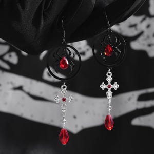 May include: A pair of silver earrings with a black spider and red gemstone charm, and a silver cross with a red gemstone charm.
