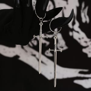 Puede incluir: Un par de pendientes de espada plateados con cierre de aro. Las espadas tienen diseños detallados y cuelgan de los aros.