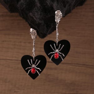 Puede incluir: Pendientes plateados con diseño de calavera y colgantes de corazón negro. Cada corazón presenta una araña plateada con una gema roja en el centro. Los pendientes se muestran sobre un fondo oscuro.