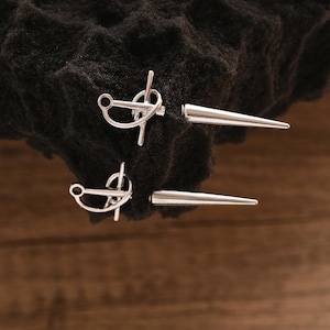 May include: Two silver-colored metal earrings with a post and a pointed end. The earrings are displayed on a black, textured surface against a wooden background. The earrings have a unique design.