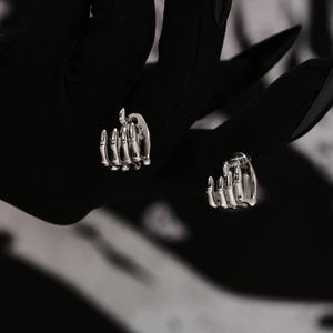 Può includere: Un paio di orecchini a forma di mano scheletrica in argento. Gli orecchini hanno la forma di una mano con le dita ricurve.