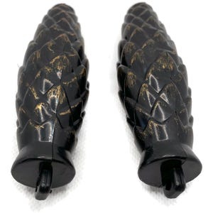 May include: Two black and gold pine cone finials with a distressed finish. The finials are approximately 10 cm long and feature a detailed design.