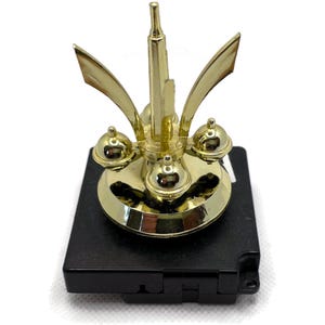 May include: Gold-toned metal clock movement with a decorative top featuring a spire and three orbs. The movement is mounted on a black plastic base.