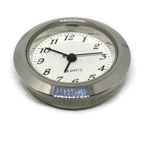May include: A silver-toned clock face with black numbers and hands. The clock face is surrounded by a silver-toned bezel and a clear glass cover. The clock face reads "QUARTZ".