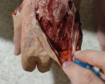 May include: A close-up of a hand with a large, open wound. The skin is torn and bloody, revealing the underlying muscle and tissue. The wound is very realistic and appears to be made of latex or silicone.