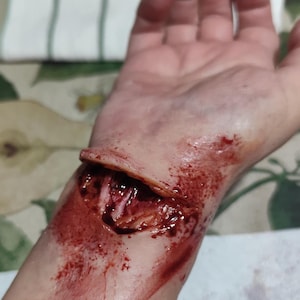 May include: A close-up of a fake, bloody wound on a person's wrist. The wound is open and appears to be deep, with red and brown makeup simulating blood.