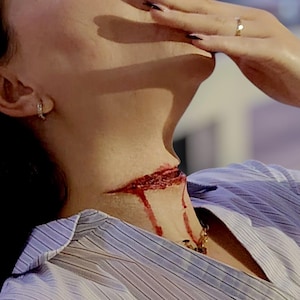 Może przedstawiać: Osoba w jasnoniebieskiej koszuli w paski, z fałszywą raną na szyi. Trzyma zapalonego papierosa, patrząc w górę. Na nadgarstku widoczny jest zegarek.