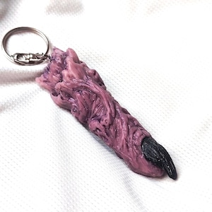 Könnte beinhalten: Ein Schlüsselanhänger in Form eines modellierten, strukturierten, malvenfarbenen Fingers mit einem markanten, gebogenen, schwarzen Fingernagel. Der Finger hat eine detaillierte, faltige Oberflächenstruktur und ist an einem silberfarbenen Metallring befestigt.