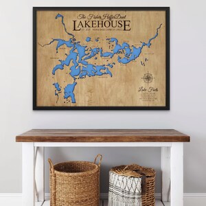 May include: Framed wooden map of a lake with blue water features. The map is labeled "Lakehouse" with the text "The Fisher Hefter Dant" above it. A compass rose and the words "Lake Facts" are also visible. The frame is black.