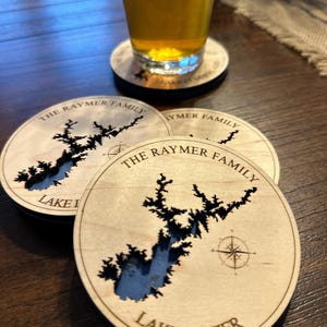 May include: Wooden coasters with a laser-cut map of Lake Lanier, featuring the text "THE RAYMER FAMILY" and "LAKE LANIER". A glass of beer sits on a coaster. The coasters are light brown with a dark brown lake design.