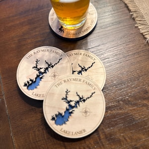 Puede incluir: Tres posavasos de madera con contornos de lagos cortados con l&aacute;ser y el texto "The Raymer Family" y los nombres de diferentes lagos. Los posavasos est&aacute;n sobre una mesa de madera.