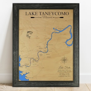 Puede incluir: Un mapa de madera del lago Taneycomo en Misuri, con un contorno azul del lago y una rosa de los vientos. El mapa incluye el texto "Lake Taneycomo, Missouri" y "Lake Facts" con detalles sobre el &aacute;rea de la superficie del lago, la longitud de la costa, la elevaci&oacute;n, la profundidad m&aacute;xima y el a&ntilde;o de finalizaci&oacute;n.