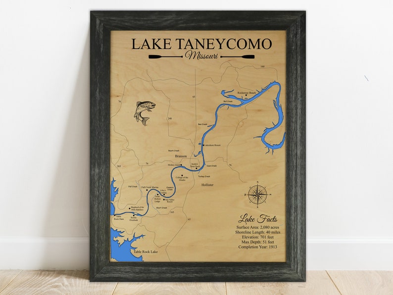 Puede incluir: Un mapa de madera del lago Taneycomo en Misuri, con un contorno azul del lago y una rosa de los vientos. El mapa incluye el texto "Lake Taneycomo, Missouri" y "Lake Facts" con detalles sobre el &aacute;rea de la superficie del lago, la longitud de la costa, la elevaci&oacute;n, la profundidad m&aacute;xima y el a&ntilde;o de finalizaci&oacute;n.