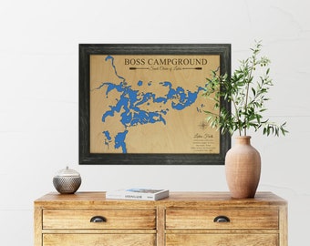 May include: A framed wooden map of Boss Campground in the South Chain of Lakes. The map is in a black frame and features a blue outline of the lakes. The map is on a wooden table with a vase of greenery and a decorative bowl.