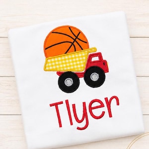 Op de afbeelding: Een wit t-shirt met een geborduurde afbeelding van een rode kiepwagen die een grote oranje basketbal in de laadbak vervoert. De vrachtwagen heeft gele ruitjesdetails aan de zijkant. De naam Tlyer is in het rood onder de afbeelding geborduurd.