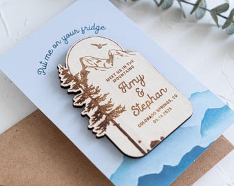 May include: A wooden magnet with a mountain and tree design. The magnet has the names "Amy & Stephan" and the date "09.16.2025" engraved. The text "Meet us in the mountains" is also present. The magnet is on a light blue card with the text "Put me on your fridge".