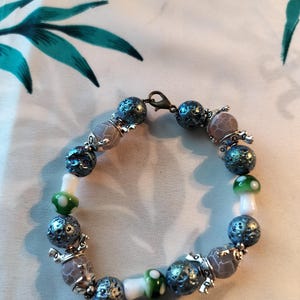 May include: A beaded bracelet featuring a variety of beads in shades of blue, gray, white, and green. The bracelet includes textured blue beads, gray stone-like beads, white beads, and green mushroom-shaped beads. The bracelet is secured with a bronze-colored clasp.