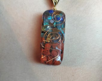 May include: A rectangular pendant featuring embedded red, blue, and turquoise stones encased in clear resin. The piece is wrapped in decorative copper wire and suspended from a black cord by a gold-toned metal bail with an etched pattern.