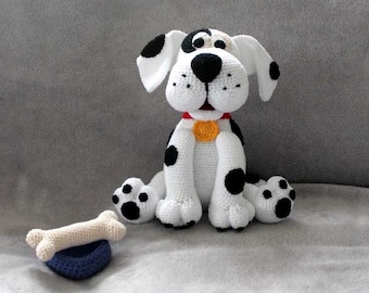 May include: A crocheted dalmatian dog plush toy with black spots, white fur, and a red collar with a yellow button. The dog is sitting on a gray surface with a white bone toy in front of it.
