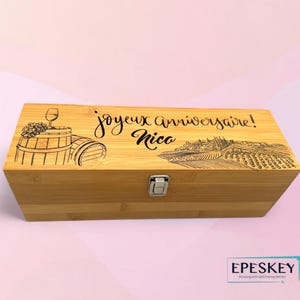 May include: A rectangular bamboo wine box with a silver clasp. The lid is engraved with a wine barrel, grapes, a wine glass, and the words "joyeux anniversaire! Nico" and a vineyard scene. The box is a light brown color.