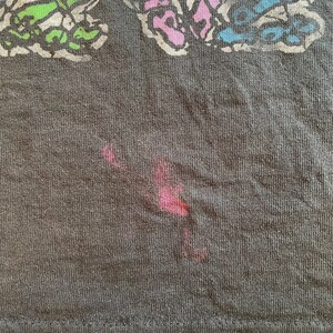 K&ouml;nnte beinhalten: Ein dunkelgraues T-Shirt mit einem bunten Schmetterlingsdesign. Der Schmetterling besteht aus verschiedenen farbigen Linien und Formen. Auf dem Shirt befindet sich ein roter Fleck.