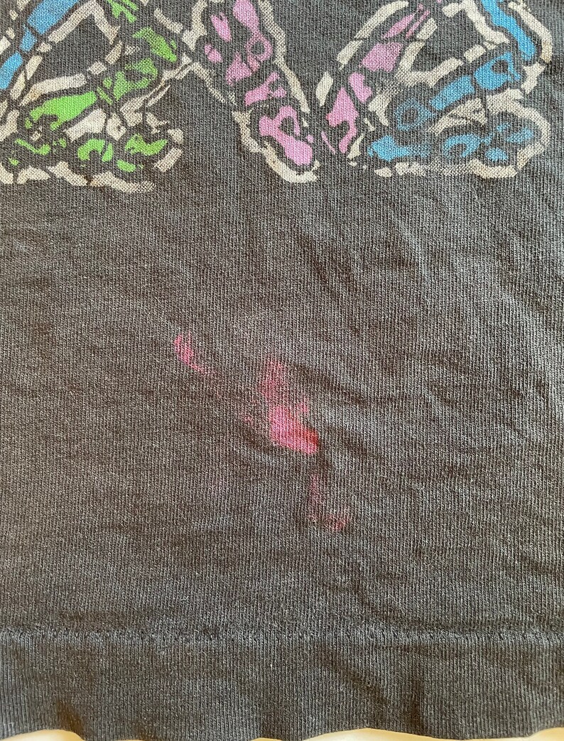 K&ouml;nnte beinhalten: Ein dunkelgraues T-Shirt mit einem bunten Schmetterlingsdesign. Der Schmetterling besteht aus verschiedenen farbigen Linien und Formen. Auf dem Shirt befindet sich ein roter Fleck.