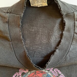 K&ouml;nnte beinhalten: Ein schwarzes T-Shirt mit einem verblassten Grateful Dead-Logo auf der Vorderseite. Das Logo ist ein bunter Wirbel mit roten, blauen und wei&szlig;en Akzenten. Das Shirt hat einen abgenutzten, Vintage-Look mit einem ausgefransten Ausschnitt.