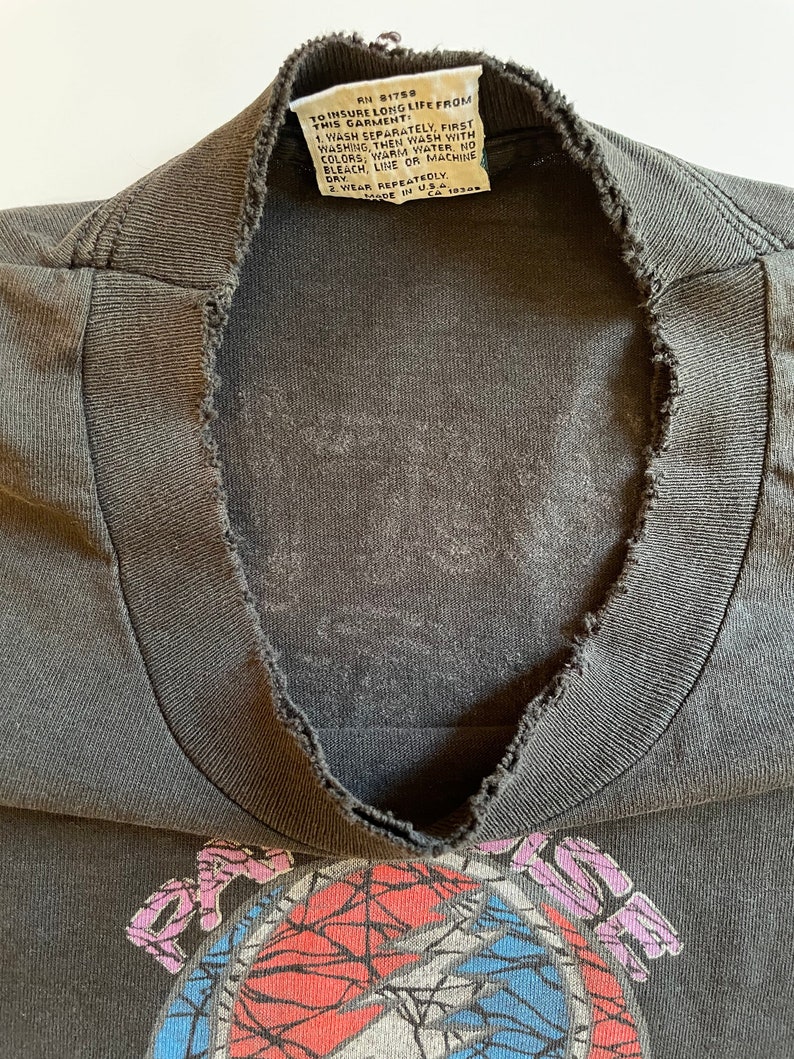 K&ouml;nnte beinhalten: Ein schwarzes T-Shirt mit einem verblassten Grateful Dead-Logo auf der Vorderseite. Das Logo ist ein bunter Wirbel mit roten, blauen und wei&szlig;en Akzenten. Das Shirt hat einen abgenutzten, Vintage-Look mit einem ausgefransten Ausschnitt.