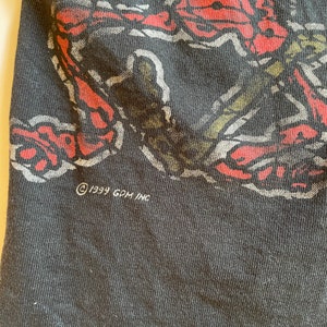 K&ouml;nnte beinhalten: Schwarzes T-Shirt mit einem roten und wei&szlig;en grafischen Aufdruck eines Schmetterlings. Das Shirt hat den Text "&copy; 1999 GDM INC" unten aufgedruckt.