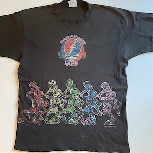 K&ouml;nnte beinhalten: Schwarzes T-Shirt mit einem bunten Grateful Dead "Steal Your Face" Logo und dem Text "Paradise Waits". Der untere Teil des Shirts zeigt eine Reihe tanzender Skelette in verschiedenen Farben.