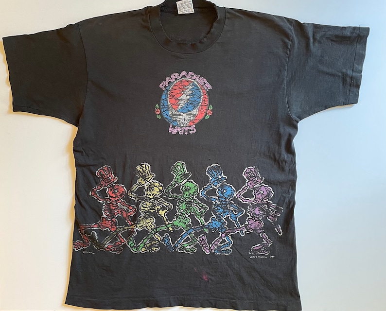 K&ouml;nnte beinhalten: Schwarzes T-Shirt mit einem bunten Grateful Dead "Steal Your Face" Logo und dem Text "Paradise Waits". Der untere Teil des Shirts zeigt eine Reihe tanzender Skelette in verschiedenen Farben.