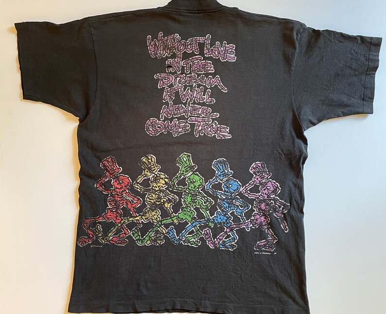 K&ouml;nnte beinhalten: Schwarzes T-Shirt mit einem Aufdruck von tanzenden Skeletten, die Zylinder tragen. Der Text "Without Love in the Dream It Will Never Come True" ist auf dem Shirt aufgedruckt.