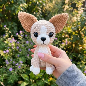 May include: A crocheted Chihuahua dog with tan and white yarn. The dog has large, tan ears, a white chest, and black eyes. The handmade dog is held in a person's hand against a background of green foliage and purple flowers.