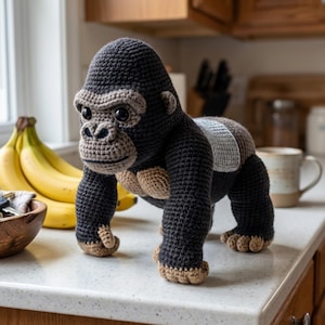 May include: A handmade crochet gorilla in shades of gray, brown, and tan. The gorilla is standing on all fours, with detailed facial features and a textured body. The toy is displayed on a countertop near a bunch of bananas.