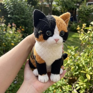 Könnte beinhalten: Handgefertigte Häkelkatzenfigur in Schildpattfarben: Schwarz, Orange und Weiß. Die Katze hat große, ausdrucksstarke Augen und eine rosa Nase. Die Häkelarbeit ist detailliert und zeigt die Textur des Garns. Ein bezaubernder Dekorationsartikel.