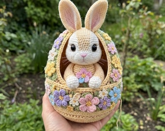Könnte beinhalten: Ein gehäkelter Osterhase in einem dekorativen Ei. Der Hase ist cremefarben mit rosa Akzenten und sitzt in einem gelben Ei, das mit bunten gehäkelten Blumen verziert ist. Das Ei wird in einer Hand gehalten.