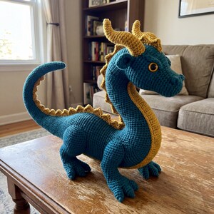 May include: A teal crochet dragon with golden yellow detailing. The dragon has a long tail, horns, and a spiky spine. It is placed on a wooden table. This handmade dragon is a whimsical decorative item.