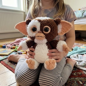 Può includere: Un peluche Gremlin fatto a mano all'uncinetto. Il giocattolo presenta una combinazione di colori bianco e marrone con grandi occhi neri, orecchie e piedi color pesca. Il peluche è tenuto in grembo da una persona.