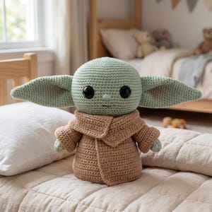 Puede incluir: Mu&ntilde;eco de ganchillo de un personaje con orejas grandes, ojos negros grandes y un abrigo marr&oacute;n. El mu&ntilde;eco es de color verde claro y marr&oacute;n, y est&aacute; sentado en una cama con una almohada blanca. El fondo incluye una estructura de cama de madera y una ventana.