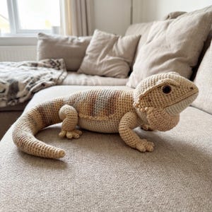 Peut inclure: Un dragon barbu crocheté aux tons beige, beige clair et marron, avec des coutures détaillées. Le reptile fait main est posé sur un canapé aux tons neutres, mettant en valeur son corps texturé et ses caractéristiques réalistes. Un objet de décoration intérieure unique et fait à la main.