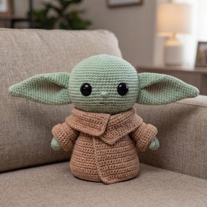 Puede incluir: Mu&ntilde;eco de ganchillo de un personaje con orejas grandes, ojos negros grandes y un abrigo marr&oacute;n. El mu&ntilde;eco es de color verde claro y marr&oacute;n, con un patr&oacute;n de ganchillo detallado. El mu&ntilde;eco est&aacute; sentado en un sof&aacute; beige.