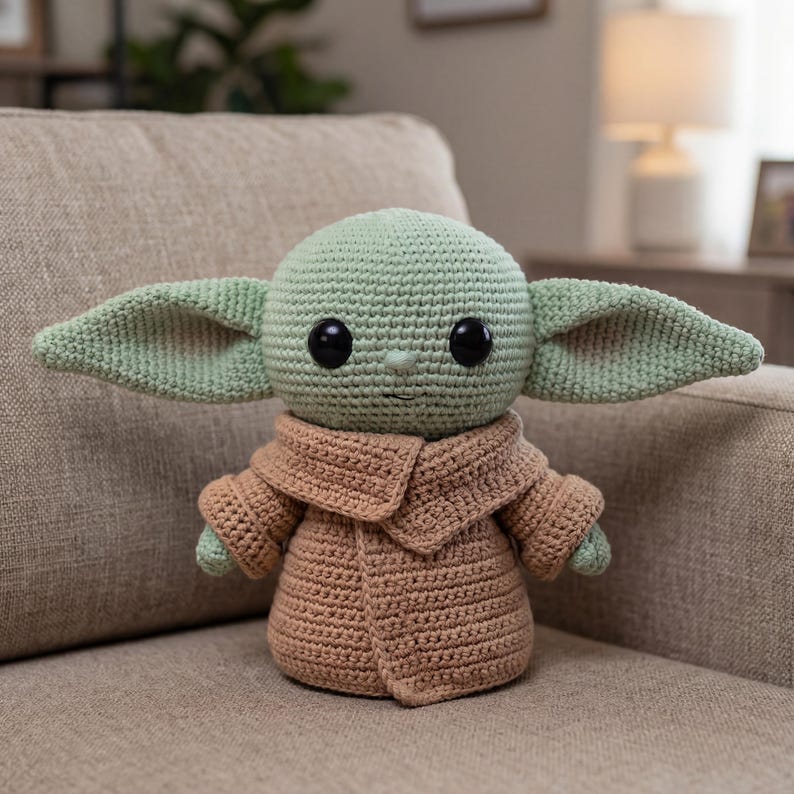 Puede incluir: Mu&ntilde;eco de ganchillo de un personaje con orejas grandes, ojos negros grandes y un abrigo marr&oacute;n. El mu&ntilde;eco es de color verde claro y marr&oacute;n, con un patr&oacute;n de ganchillo detallado. El mu&ntilde;eco est&aacute; sentado en un sof&aacute; beige.