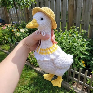 Könnte beinhalten: Eine handgefertigte weiße Häkelgans mit gelbem Hut und gelb-weiß kariertem Kragen. Die Gans hat gelbe Füße und einen gelben Schnabel. Die Gans wird vor einem Holzzaun und grünem Laub gehalten.